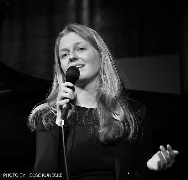 The black and white photo shows a young woman with long blonde hair singing into a microphone and smiling. She is wearing a dark-coloured top and is obviously performing live on stage.