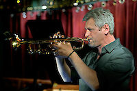 A jazz musician with grey hair concentrates on playing the trumpet on a stage with a red curtain in the background. The scene shows a typical jazz club atmosphere with warm lighting and blurred points of light in the background.