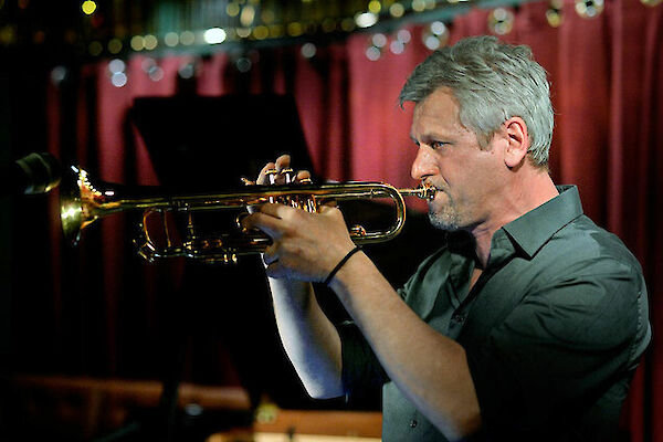 A jazz musician with grey hair concentrates on playing the trumpet on a stage with a red curtain in the background. The scene shows a typical jazz club atmosphere with warm lighting and blurred points of light in the background.