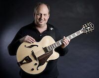 Ein Musiker lächelt. Er trägt ein schwarzes Hemd. Er hält eine helle Jazz-Gitarre. Die Gitarre ist hohl. Im Hintergrund ist eine graue Wand. Das ist ein Studio. Das Foto zeigt den Gitarristen. Er steht in einer guten Pose. Er zeigt seine Gitarre.