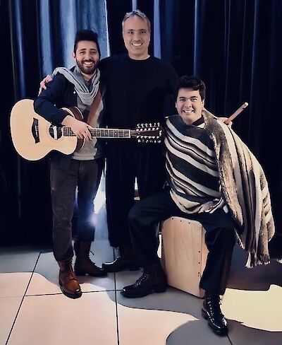 The picture shows three smiling men in a studio with a dark background, posing as a music group. On the left is a man with a guitar, in the centre is a man in a black jumper and on the right is a man with a striped poncho sitting on a cajon (percussion instrument).