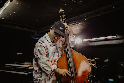 The picture shows a young musician wearing a baseball cap, playing a double bass with concentration and looking slightly downwards. The picture was obviously taken during a live performance in a club or concert hall, recognisable by the atmospheric lighting and the stage equipment in the background.