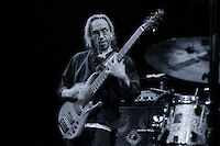 Das Foto ist schwarz-weiß. Es zeigt einen Musiker. Der Musiker hat lange Haare. Er trägt eine Brille. Er spielt E-Bass. Er ist sehr konzentriert. Im Hintergrund sieht man ein Schlagzeug. Man sieht auch Verstärker. Das ist eine Live-Show oder eine Studio-Aufnahme.