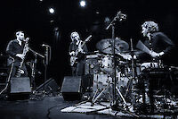 Das Schwarz-Weiß-Foto zeigt drei Musiker während einer Live-Performance auf einer Bühne: einen Saxophonisten links, einen Bassisten in der Mitte und einen Schlagzeuger rechts. Die atmosphärische Beleuchtung und die professionelle Bühnenausstattung mit Mikrofonen und Verstärkern unterstreichen den konzertanten Charakter der Aufnahme.