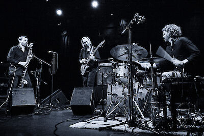 Das Foto ist schwarz-weiß. Es zeigt drei Musiker auf einer Bühne. Links spielt ein Mann Saxophon. In der Mitte spielt ein Mann Bass. Rechts spielt ein Mann Schlagzeug. Die Musiker geben ein Konzert. Es gibt Mikrofone und Verstärker auf der Bühne. Das Licht macht eine schöne Stimmung.