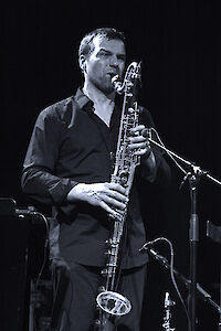 Das ist ein Schwarz-Weiß-Foto. Es zeigt einen Mann. Der Mann spielt Saxophon. Er trägt ein dunkles Hemd. Er spielt sehr konzentriert. Das Licht ist dramatisch. Der Hintergrund ist schwarz. Man sieht ein Mikrofon. Der Mann spielt live vor Publikum.