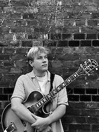 Das schwarz-weiße Foto zeigt einen jungen Mann mit blonden Haaren in einem gestreiften Hemd, der eine E-Gitarre hält. Er sitzt vor einer alten, verwitterten Backsteinmauer und blickt nachdenklich zur Seite.