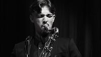 Das schwarz-weiße Foto zeigt einen Saxophonisten mit gewellten Haaren und Sonnenbrille, der intensiv in sein Instrument spielt. Die dramatische Beleuchtung vor dunklem Hintergrund verleiht der Aufnahme eine atmosphärische, künstlerische Wirkung.