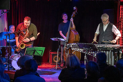 In einem Jazz-Club spielen vier Musiker. Es gibt einen Schlagzeuger. Es gibt einen Saxophon-Spieler. Es gibt einen Bass-Spieler. Es gibt einen Vibraphon-Spieler. Die Bühne hat rotes Licht. Die Bühne hat blaues Licht. Vorne sitzen Zuschauer. Man sieht ihre Schatten.