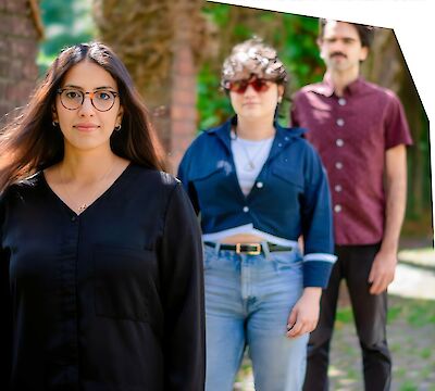 Das Bild zeigt drei Personen in legerer Kleidung, die in einem Park oder auf einem Universitätscampus stehen, wobei eine junge Frau mit Brille und schwarzer Bluse im Vordergrund zu sehen ist. Im Hintergrund befinden sich zwei weitere Personen - eine Frau in blauer Bluse und Jeans sowie ein Mann in bordeauxrotem Hemd - vor grüner Vegetation und Backsteingebäuden.