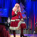 Ein Mann trägt ein rotes Weihnachts-Kostüm. Er hat eine rosa Perücke auf. Er hat auch eine Weihnachts-Mütze auf. Er steht auf einer Bühne. Er hat ein Mikrofon in der Hand. Er macht eine Show. Die Bühne ist schön geschmückt. Es gibt blaues Licht. Es gibt Vorhänge. Es gibt Weihnachts-Schmuck. Im Hintergrund steht ein Klavier.