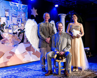 The picture shows three actors on a theatre stage with a Christmas stage set, including two men in grey suits and a woman in a golden dress. White angel wings, a star and a backdrop of oriental buildings can be seen in the background, alluding to a Christmas story.