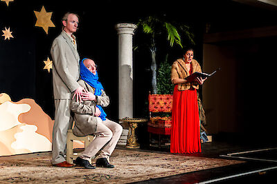 The picture shows three actors on a theatre stage, with one man standing in a grey suit, a second man sitting in a blue headscarf and a woman in a red dress reading from a book. The stage is decorated with golden stars, an antique column and oriental props as a backdrop for a Christmas performance.