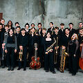 The picture shows a group of young musicians in black clothing posing with their instruments in front of a grey concrete wall. The big band consists of around 20 people with various jazz instruments such as saxophones, trumpets, trombones, guitar, double bass and drums.