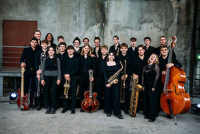 The picture shows a group of young musicians in black clothing posing with their instruments in front of a grey concrete wall. The big band consists of around 20 people with various jazz instruments such as saxophones, trumpets, trombones, guitar, double bass and drums.