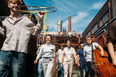 The picture shows a group of jazz musicians with their instruments in front of an industrial backdrop with brick buildings and chimneys in bright sunshine. A young man with a trombone can be seen in the foreground, while other musicians with saxophones, double basses and other instruments stand in the background.