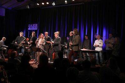 The picture shows a jazz performance at the Jazz-Schmiede with a large group of musicians on stage playing various instruments such as saxophones, trumpets and keyboards. The illuminated "Jazz-Schmiede" logo can be seen in the background, while the audience follows the live performance in the dark auditorium.