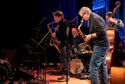 The picture shows a live jazz performance at the Jazz-Schmiede with several musicians on stage, including saxophonists in the foreground and a drummer and a double bass player in the background. The musicians can be seen in an intimate club atmosphere with blue lighting and the characteristic Jazz-Schmiede logo in the background.
