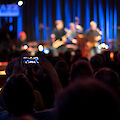 The image shows a concert scene in a club or small concert hall, with a spectator filming the performance with his smartphone in the foreground. In the background, musicians on a blue-lit stage can be recognised out of focus, while the audience follows the performance in the dark auditorium.