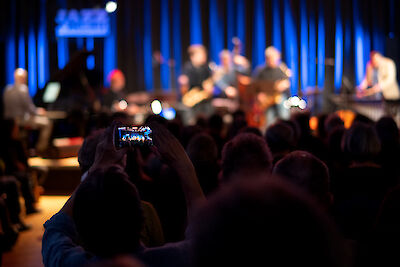 The image shows a concert scene in a club or small concert hall, with a spectator filming the performance with his smartphone in the foreground. In the background, musicians on a blue-lit stage can be recognised out of focus, while the audience follows the performance in the dark auditorium.