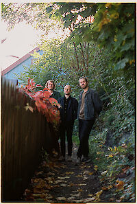 Das Bild zeigt drei Männer. Sie stehen auf einem Weg im Wald. Der Weg ist schmal. Um sie herum sind viele Pflanzen. Die Blätter sind bunt. Es ist Herbst. Im Hintergrund steht ein blaues Haus. Die Sonne scheint. Das macht eine schöne Stimmung.