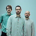 Das Bild zeigt drei Männer. Sie stehen vor einem blauen Hintergrund. Alle tragen helle Hemden. Der Mann in der Mitte steht vorne. Er trägt ein grünes Hemd. Links steht ein Mann mit Brille. Er trägt ein blaues Hemd. Rechts steht ein Mann ohne Haare. Er hat einen Bart. Er trägt ein graues Hemd.