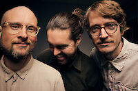 Das Bild zeigt drei Männer. Sie sind ganz nah zu sehen. Der Hintergrund ist dunkel. Zwei Männer tragen eine Brille. Alle drei schauen freundlich. Der Mann in der Mitte hat lange Haare. Seine Haare sind zu einem Knoten gebunden. Er schaut etwas nach unten. Die anderen zwei Männer tragen helle Hemden. Sie schauen direkt in die Kamera.