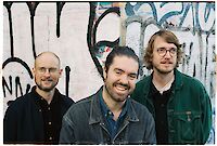 Das Bild zeigt drei Männer. Sie stehen vor einer bunten Wand. Die Wand hat Graffiti. Die Männer lächeln in die Kamera. Links steht ein Mann mit Glatze. Er trägt eine Brille. Er hat ein schwarzes Hemd an. In der Mitte steht ein Mann mit dunklen Haaren. Er trägt eine Jeans-Jacke. Rechts steht ein Mann mit lockigen Haaren. Er trägt auch eine Brille. Er hat eine grüne Jacke an.