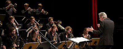 Das Bild zeigt eine Big Band während eines Auftritts mit einem Dirigenten im Vordergrund, der das Ensemble mit ausgestreckten Händen leitet. Die Musiker sitzen in typischer Big Band-Formation mit Blechbläsern in den hinteren Reihen und Holzbläsern sowie Rhythmusgruppe im vorderen Bereich, alle in dunkler Kleidung auf einer Bühne mit rotem Vorhang im Hintergrund.