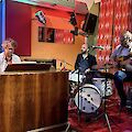 Das Bild zeigt eine Jazz-Band. Es sind drei Musiker. Sie spielen in einem Club. Der Club hat schönes Licht.

Links sitzt ein Mann am Piano. Das Piano ist aus Holz. Der Mann hat lockige Haare. Er trägt eine Brille.

In der Mitte spielt ein Mann Schlagzeug.

Rechts sitzt ein Mann auf einem Hocker. Er spielt Gitarre. Er hat graue Haare.