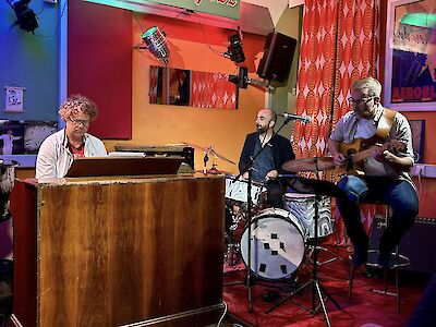 The picture shows a jazz band with three musicians performing in an atmospherically lit club or bar. On the left is a pianist with curly hair and glasses sitting at a wooden piano, in the middle is a drummer playing the drum set and on the right is a guitarist with grey hair sitting on a stool.