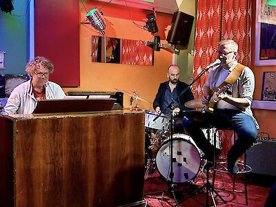 The picture shows three musicians during a live performance in a club with colourful lighting and decorative wall elements. From left to right, a pianist on a wooden piano, a drummer on a drum set and a guitarist with glasses can be seen making music together in a relaxed atmosphere.