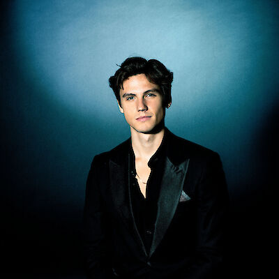 Das Bild zeigt ein professionelles Porträt eines jungen Mannes mit dunklen, gewellten Haaren, der elegant in einem schwarzen Anzug mit offenem Hemd gekleidet ist. Das Foto ist vor einem bläulich beleuchteten Hintergrund aufgenommen und wirkt wie eine hochwertige Studiofotografie oder ein Pressefoto.