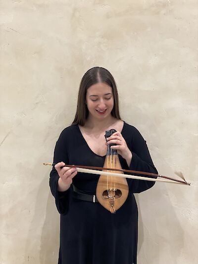 Das Bild zeigt eine lächelnde Musikerin in einem schwarzen Kleid, die vor einer hellen Wand steht und ein traditionelles Streichinstrument mit einem langen Bogen spielt. Das Instrument hat einen charakteristischen dreieckigen Holzkorpus mit Saiten und wird mit einem Streichbogen gespielt.