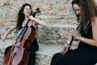 Zwei Frauen sitzen vor einer alten Mauer aus Stein. Sie tragen schwarze Kleidung. Sie spielen Musik. Eine Frau hat lange dunkle Haare. Sie spielt Cello. Die andere Frau hat lockige Haare. Sie spielt Lyra. Die Frauen sind konzentriert. Die alte Steinmauer sieht verwittert aus.