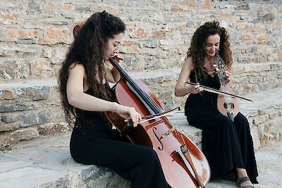 Das Bild zeigt zwei Musikerinnen in schwarzer Kleidung, die auf Steinstufen vor einer rustikalen Backsteinmauer sitzen und gemeinsam musizieren. Die linke Frau spielt Cello, während die rechte Musikerin ein Streichinstrument mit einem Bogen spielt.