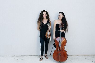 Zwei Frauen lachen. Sie haben schwarze Kleidung an. Sie stehen vor einer weißen Wand. Sie haben Musik-Instrumente. Die linke Frau hat lockige Haare. Sie hält eine Lyra. Die rechte Frau hat lange dunkle Haare. Sie steht neben einem Cello.
