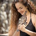 Das Bild zeigt eine lächelnde Frau mit lockigen braunen Haaren in einem schwarzen Oberteil, die ein traditionelles Saiteninstrument mit vielen Saiten spielt. Sie hält das Instrument liebevoll in den Händen und scheint konzentriert zu musizieren, während der Hintergrund unscharf verschwommen ist.