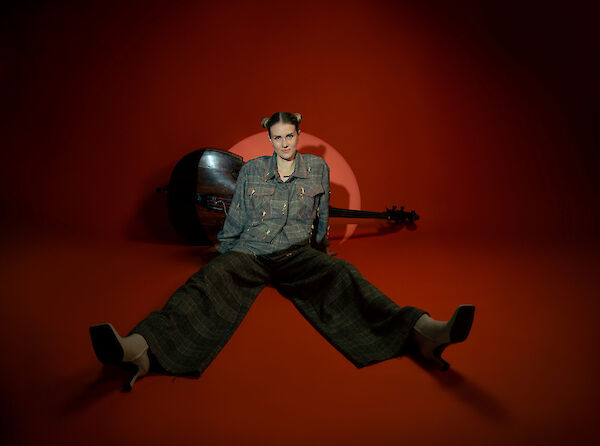 A woman in grey clothing sits on the floor with her legs apart in front of a reddish background, holding a double bass. The bassist is wearing a jacket and wide trousers, while the large black bass is positioned diagonally behind her.