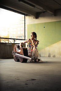 A woman in a colourful dress adorned with flowers sits next to her double bass in an industrial underground car park or multi-storey car park. The sunlight falls through a large window and illuminates the musician in the urban concrete environment.