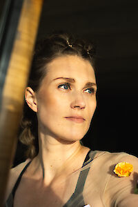 A young woman with pinned-up hair and a nose ring looks thoughtfully to the side while warm sunlight illuminates her face. She wears a beige jacket with a yellow flower as an accessory and stands in front of a dark background.