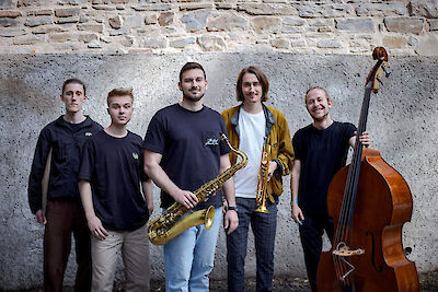 Das Bild zeigt fünf junge Männer, die als Jazz-Quintett vor einer alten Steinmauer posieren. Von links nach rechts sind ein Schlagzeuger, ein Pianist, ein Saxophonist mit seinem Instrument, ein Trompeter mit goldener Trompete und ein Kontrabassist mit seinem Instrument zu sehen.