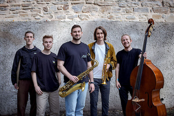 The picture shows five young men posing as a jazz quintet in front of an old stone wall. From left to right, you can see a drummer, a pianist, a saxophonist with his instrument, a trumpet player with a golden trumpet and a double bass player with his instrument.