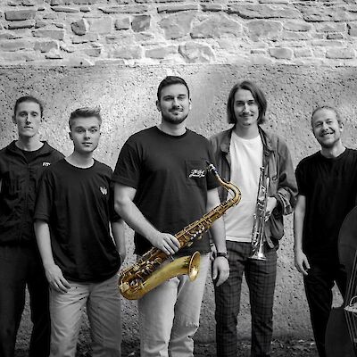 Auf dem Bild ist eine fünfköpfige Musikband zu sehen, die vor einer alten Steinmauer posiert. Die Musiker halten verschiedene Instrumente, darunter ein goldenes Saxophon und eine Trompete, wobei das Foto größtenteils in Schwarz-Weiß gehalten ist und nur die Blasinstrumente in ihrer natürlichen goldenen Farbe hervorgehoben sind.