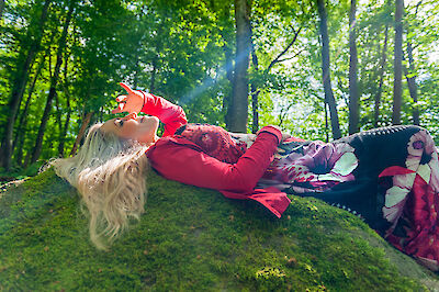 Drei Menschen liegen auf dem Boden im Wald. Der Boden hat Moos. Die Sonne scheint durch die Bäume. Die Menschen tragen rote Kleidung. Sie haben auch Kleidung mit Mustern an. Sie fühlen sich gut. Sie mögen die Natur. Das warme Licht kommt durch die grünen Blätter der Bäume.