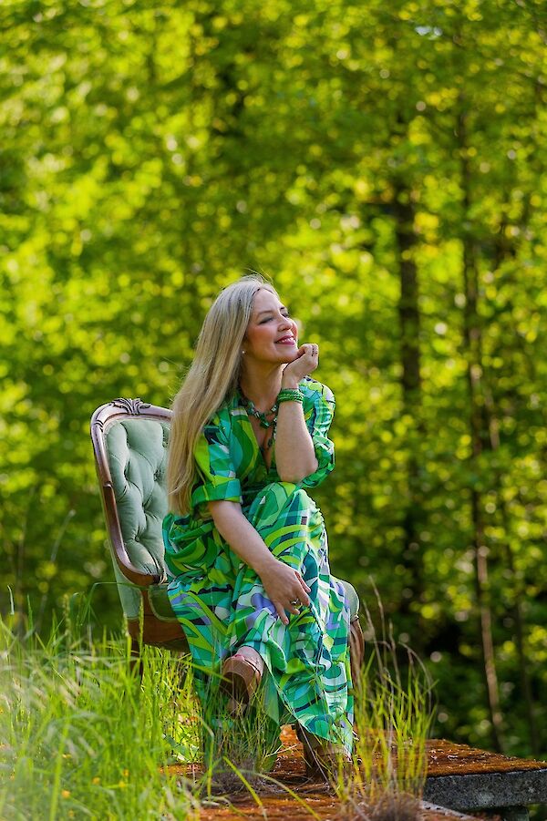 A blonde woman in a green patterned dress sits relaxed in an antique armchair in a natural park landscape. She has her eyes closed, smiles peacefully and is visibly enjoying the sun's rays against a backdrop of blurred green trees.