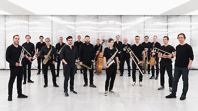 Das Bild zeigt eine Jazz-Band. Die Band hat 17 Musiker. Die Musiker tragen schwarze Kleidung. Sie stehen in einem hellen Raum. Der Raum hat weiße Wände. Der Boden hat Fliesen. Die Musiker haben verschiedene Instrumente. Sie haben Saxophone, Trompeten und Posaunen. Ein Musiker hat einen Kontrabass. Die Musiker machen ein Gruppen-Foto.