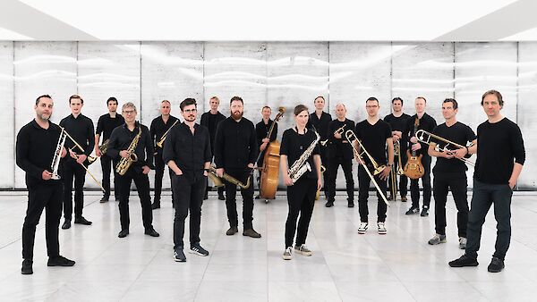 The picture shows a large jazz big band with around 17 musicians dressed in black, posing in a modern, bright room with white walls and a tiled floor. The musicians are holding various instruments such as saxophones, trumpets, trombones and a double bass and are lined up in a loose formation for a group photo.