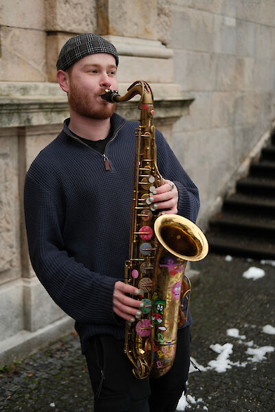 Ein junger Mann mit karierter Mütze und blauem Pullover spielt Saxophon vor einer Steinmauer. Das goldene Tenorsaxophon ist mit bunten Stickern verziert und er steht auf einem gepflasterten Boden mit Schneeresten.