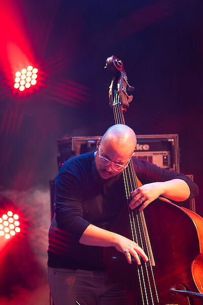 Ein Mann mit Glatze und Brille spielt konzentriert Kontrabass auf einer Bühne in dramatischem roten Bühnenlicht. Im Hintergrund sind Verstärker und helle Scheinwerfer zu sehen, die eine typische Konzert- oder Clubatmosphäre schaffen.